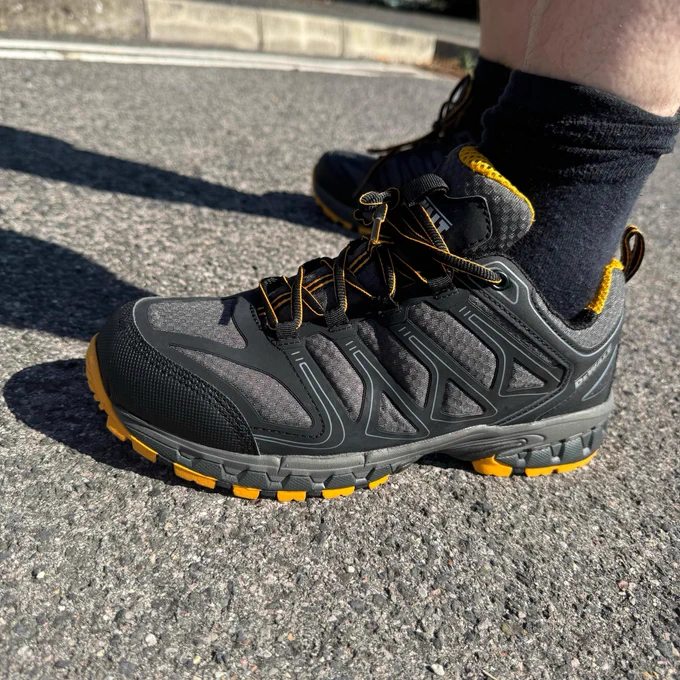 Close-up of a person wearing grey and black work shoes with yellow accents, standing on pavement.