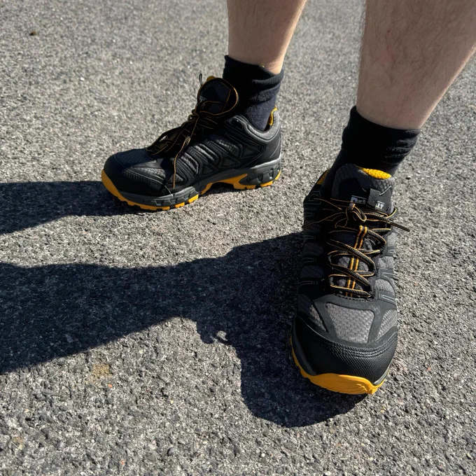 Close-up view of a person's lower legs and feet, wearing dark athletic shoes with yellow details and black socks, standing on an asphalt surface in sunlight.