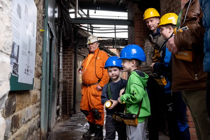 Two children wearing hard hats ready to go underground with a Mine Guide, in the background are adults stood behind them.
