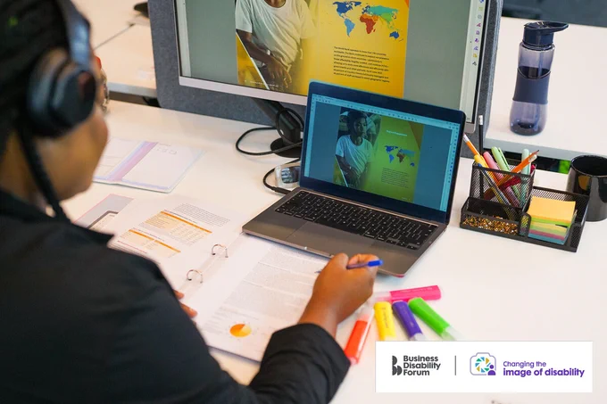 Person with less-visible disabilities works at a desk with a computer screen and laptop open in from of them. They're wearing headphones. The Business Disability Forum 'Changing the image of disability' logo is in the lower right corner of the image.