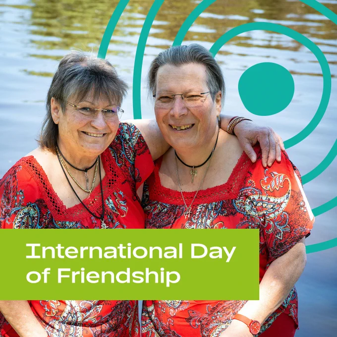 Two older women smiling at the camera, with one older woman's arm around the other's shoulders. They are wearing similar red blouses and necklaces, and standing near a body of water. The UN Decade of Healthy Ageing logo is in the background. Text: 'International Day of Friendship'.