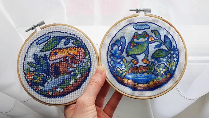 Two round embroidered hoops. On the left is a cross-stitch embroidery of a gecko in a terrarium. On the right is a cross-stitch of a frog in a terrarium.