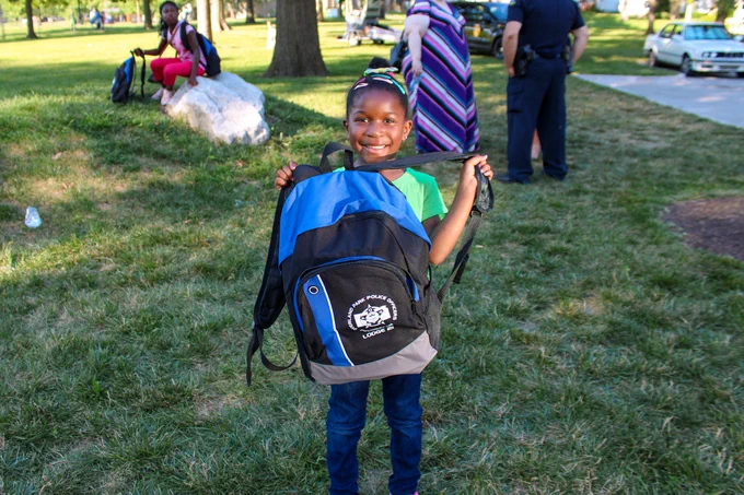 A child in a park holding up a large blue and black backpack, smiling at the camera. Other people and police officers are visible in the background.