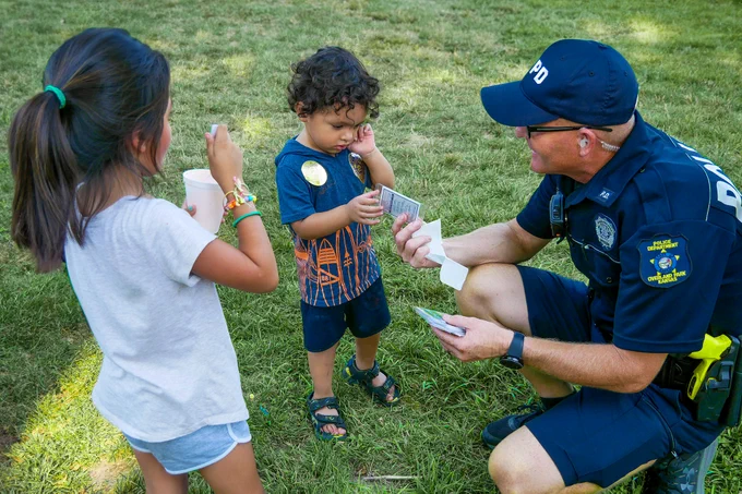 A police officer is interacting with two young children in a park, handing a badge sticker to a toddler.