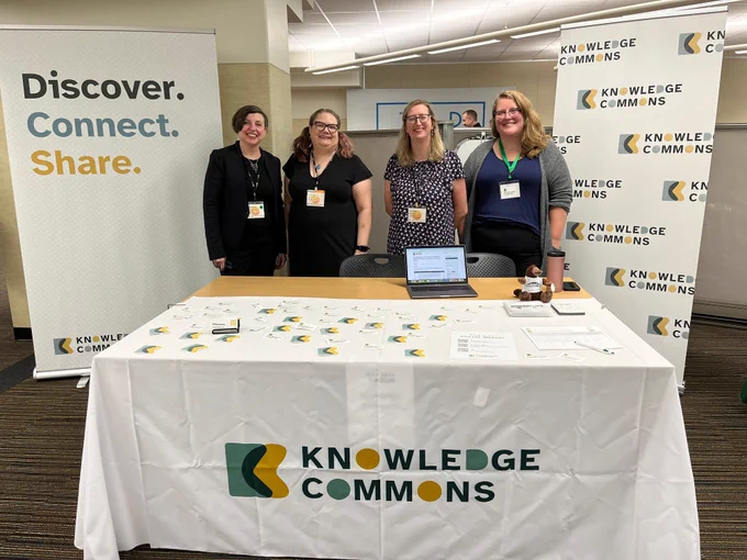 A group of women standing in front of a table with a tablecloth reading "Knowledge Commons"
