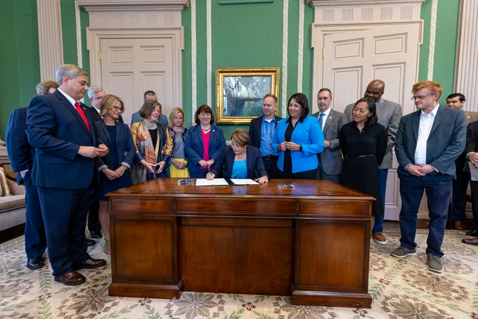 A group of people stand around a woman, Governor Healey, sitting at a large wooden desk. She is signing a document. The walls are painted green with white accents and doors. 