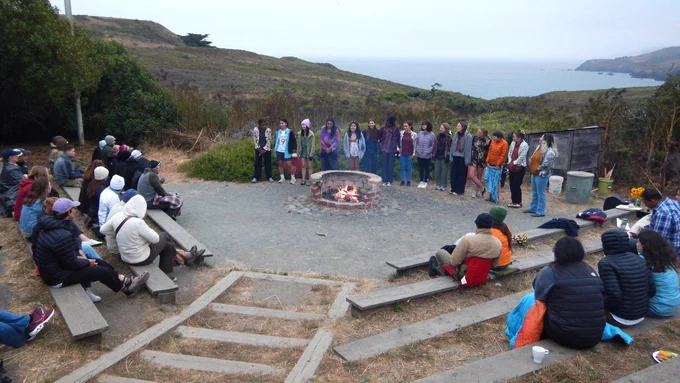 The 2024 Armstrong Scholars and their leaders stand behind a campfire lit in an outdoor amphitheater around which are seated several adults dressed in warm clothing. Hills and ocean are in the background.