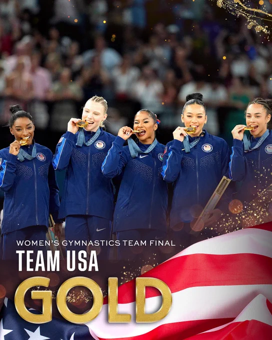Team USA at the Women's Gymnastics Team Final, smiling and celebrating with gold medals around their necks. They are wearing matching navy blue team jackets. In the background, a crowd is visible. The text at the bottom of the image reads 'TEAM USA GOLD' with an American flag motif below.