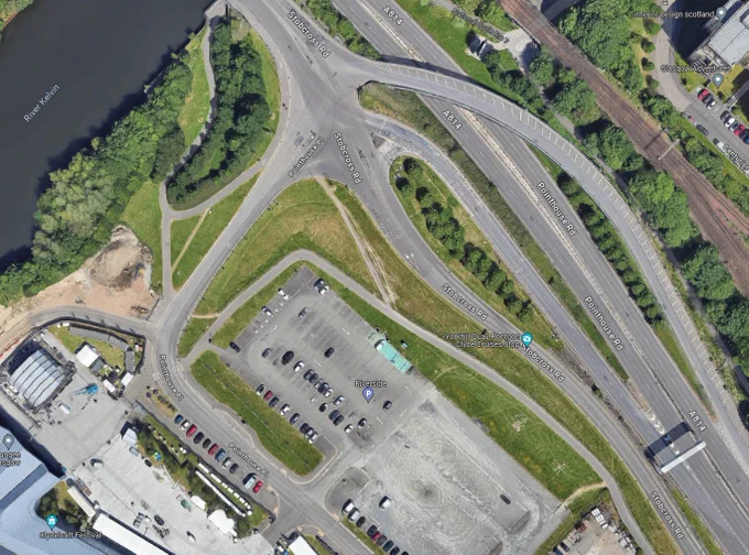 Satellite image from Google of the Transport Museum in Glasgow. A lot of the photograph is taken up with several roads and car parks. Rail lines are also visible. There are some paths in the image, but they don't take direct routes. You can see direct routes cut into the grass where people have walked direct routes.