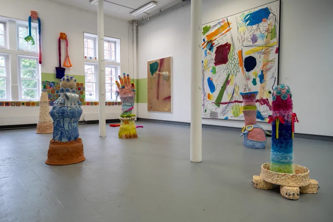Image shows a series of colourful artworks sitting in a bright white studio room. On the walls, brightly coloured paintings can be seen.