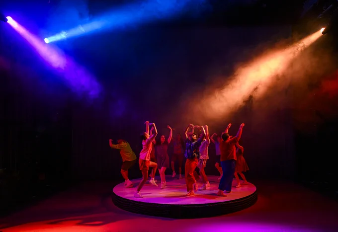 Actors perform on a round platform on a stage under purple and orange lights