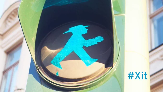 Auf dem Foto ist ein grünes Ampellicht mit dem Ostberliner Ampelmännchen zu sehen. Im Hintergrund ist ein Gebäude. Rechts unten ist steht Schriftzug #Xit.