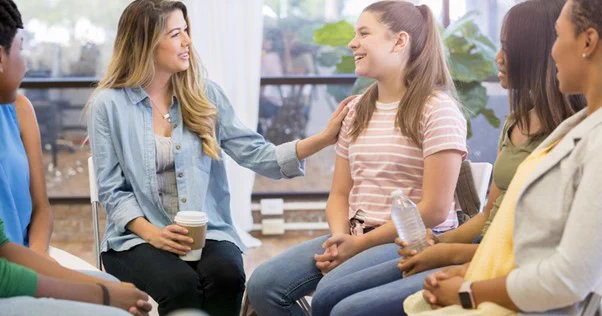 Image of woman sat on chair in a circle, a woman wearing a blue shirt has her hand on a woman's shoulder, who is wearing a pink striped top, and everyone is smiling. 