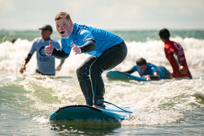 Ollie is stood on a surfboard in the sea looking happy and putting his thumbs up at the camera. He's wearing a bright blue top. Behind him are 3 others stood in the water and surfing.