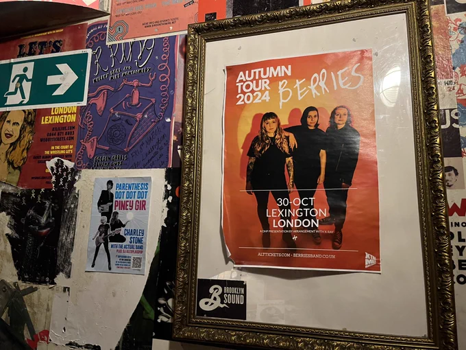 A wall cluttered with gig posters inc a prominent orange one for the BERRIES band and a very small blue one for Parenthesis Dot Dot Dot, Piney Gir and Charley Stone with the Actual Band