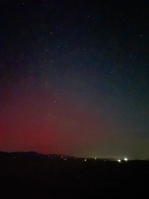 A dark, low horizon marked by a few bright lights, and a vast dark sky with recognizable constellations and barely-visible Amdromeda galaxy. The bottom left side of the image is overtaken by a smear the color of strawberry jelly, which is glowing ions in the sky.