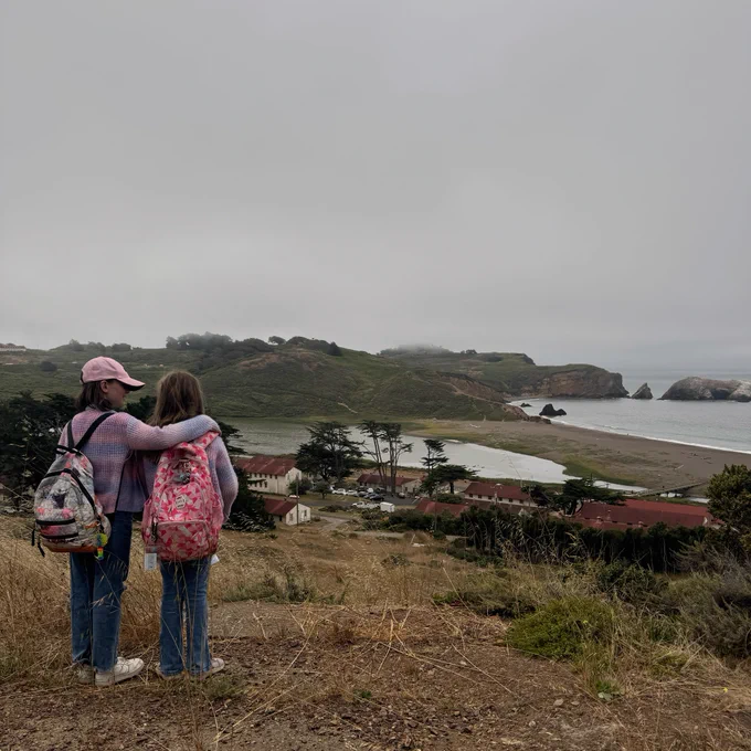 Two young campers stand with their arms around each other as they look out at a foggy view of buildings, hills, and ocean from a hilltop