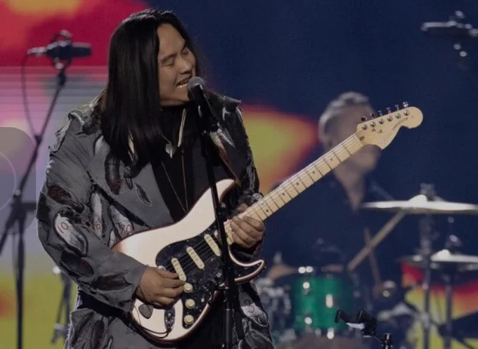 Photo of Indigenous musician Asyanabee, wearing sari coat with feathers on it, and holding a bronze electrical guitar