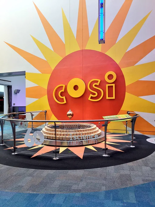COSI sign with a pendulum knocking down marbles in front of it