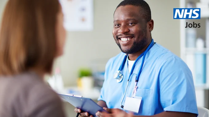 A member of staff, wearing blue scrubs and wearing a stethescope is smiling at a patient.