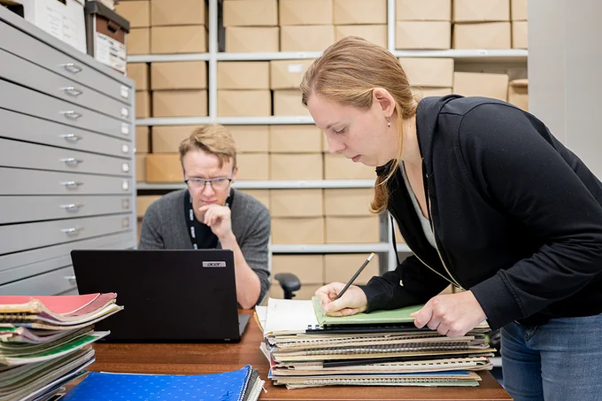 Two people at work in an archive strong room. One is leaning over a pile of papers, and the other is frowning at a laptop