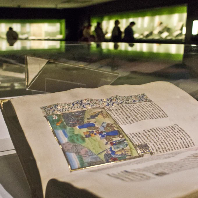 A view of a medieval book open on display with figures in an exhibition space in the background.