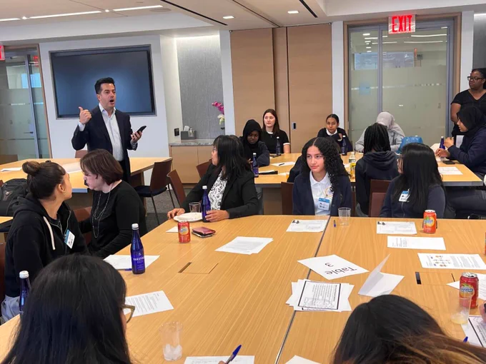 Students sit at tables with papers in front of them while listening to a presentation. 