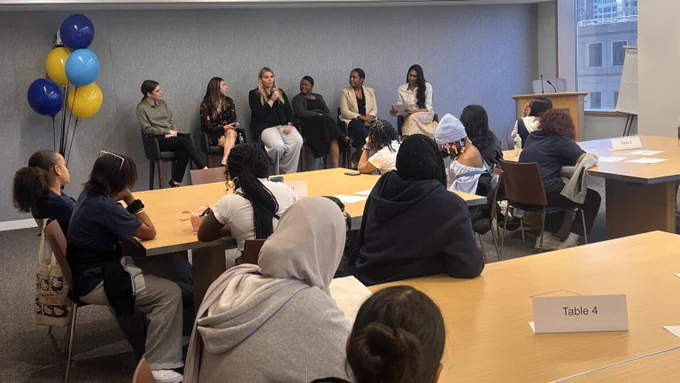 Students sit at tables and listen to a panel presentation. 