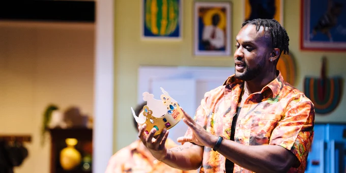 A person in a vibrant, patterned shirt is animatedly holding and looking at a decorated crown in an art-filled room.