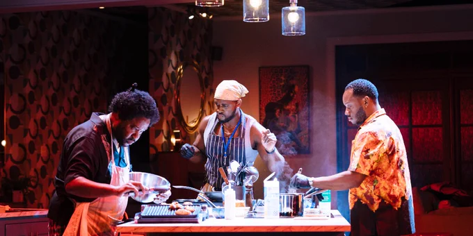Three people are actively engaged in cooking in a warmly lit kitchen decorated with hanging lights and a cozy ambiance. 