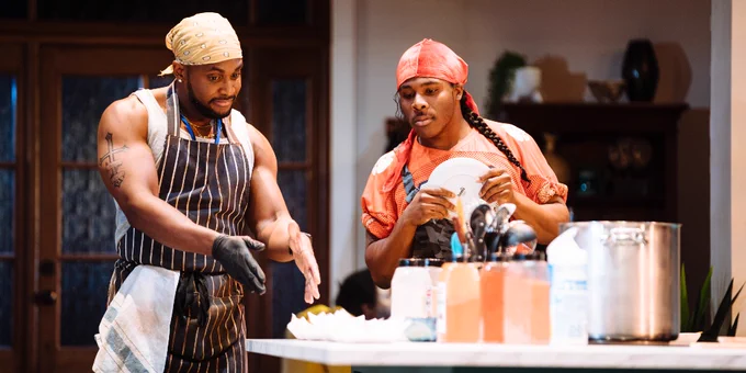 Two individuals in a kitchen setting, wearing aprons and headscarves, are focused on preparing food with various ingredients and kitchen equipment around them.