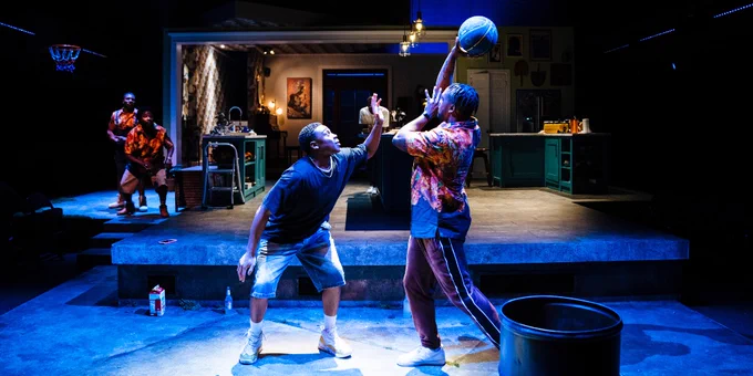 Two actors on a stage set resembling a kitchen, engaged in a playful basketball pose with one actor attempting to block the other's shot.