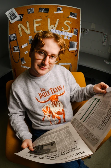 Pictured is a man sitting in front of a bulletin board that reads ‘NEWS’ with various clippings of pictures around it. The man sits on a bright orange chair and holds a newspaper with both hands while he looks into the camera. He is a redhead with short hair parted in the middle and wears light brown, circular glasses and a gray sweater that says “THE DAILY TEXAN’ in burnt orange letters with Bevo the longhorn in the middle also colored burnt orange. Photo by Manoo Sirivelu.