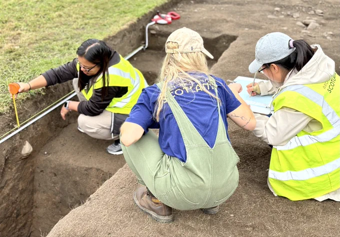 Photo of three women around an archaeological trench. One woman, to the left, has a hand tape, held against the side section of a trench. The woman on the right is drawing the section while the one in the middle, with her back to the camera, supports her.
