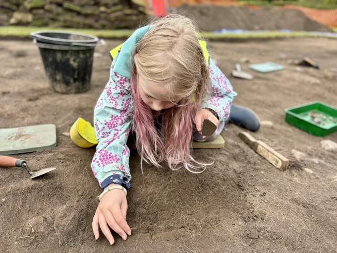 Photo of a girl in an archaeological trench, picking out an object from an area she has trowelled.
