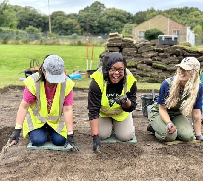 Photo of 3 women trowelling in an archaeological trench. The woman in the centre is laughing and looking into the camera.