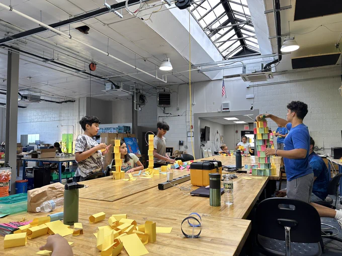 A long table is strewn with index cards in many colors . Campers and staff are building towers….