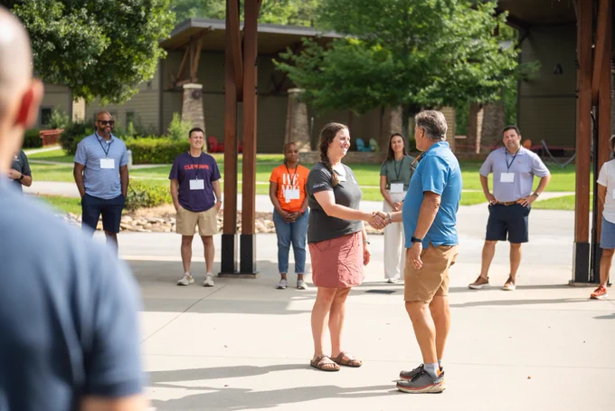 A group of people standing in a circle outdoors. Two people are in the middle of the circle shaking hands.