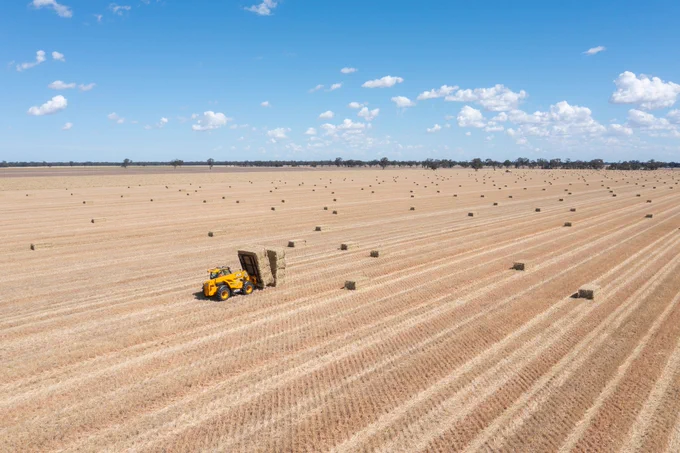 Aerial view of a vast agricultural field with multiple rectangular hay bales under a clear blue sky. A yellow tractor is actively baling hay.