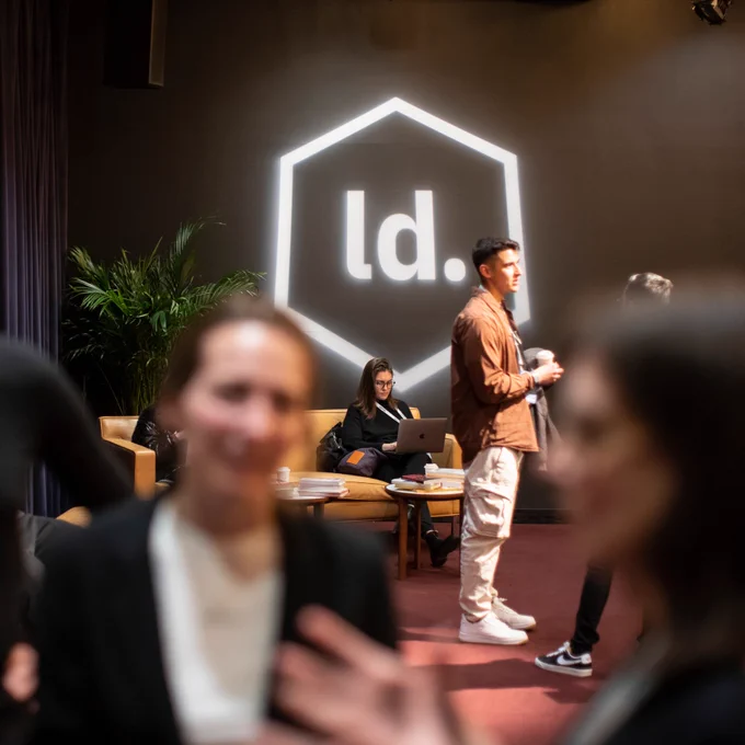 People engaged in conversations in a room, with a person working on a laptop sat on a yellow sofa in the background and an illuminated "ld." logo displayed on the wall. 