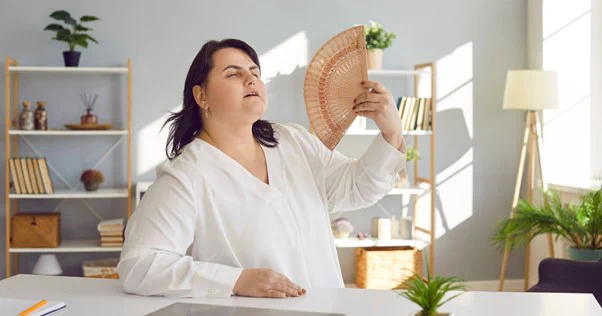 Image of woman wearing a white long sleeved top and holding a spread out hand fan in front of her face. There is a book shelf in the background, grey walls, a lamp.