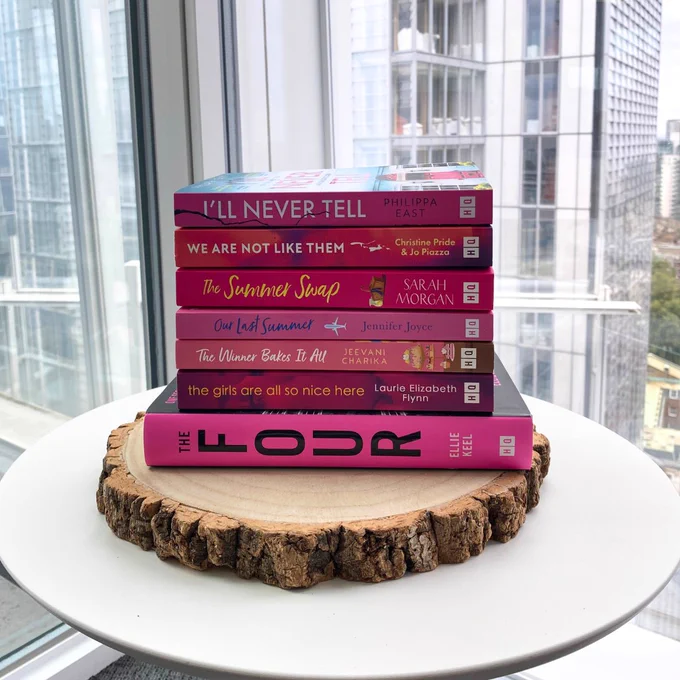 A pink stack of books on a table including I'll Never Tell, We Are Not Like Them, The Summer Swap, Our Last Summer, The Winner Bakes It All, The Girls Are All So Nice Here, The Four.