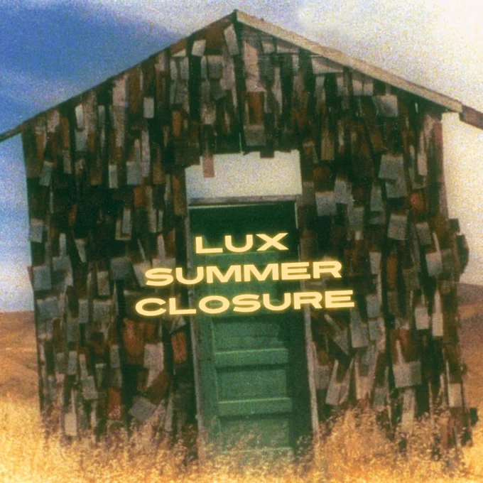A lone facade of a house with crumbling wooden tiles and a green door, slightly tilted to the left, stands against a vast, empty yellow field under a clear blue sky. 