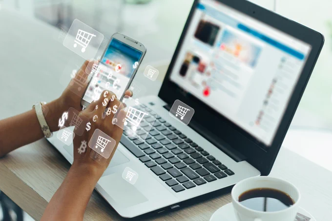 Photo of a person at a laptop. They are holding a phone with digital symbols depicting a shopping cart and currencies. 