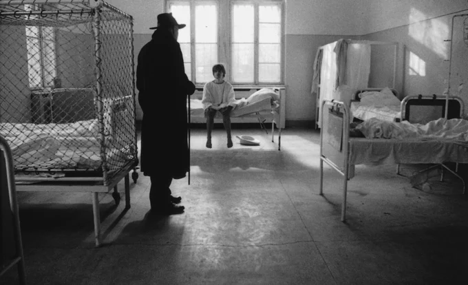Still from Werckmeister Harmonies showing man sat on hospital bed next to a netted hospital bed and a shadowy silhouette of a man with a stick in a coat and hat.