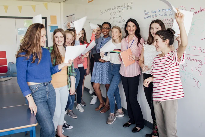 A diverse group of women smile and wave papers in the air in a classroom. 