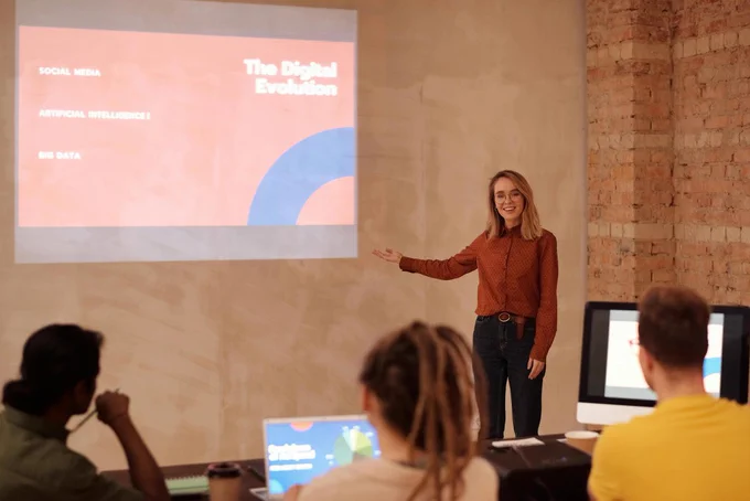 Eine weiblich gelesene Person erklärt an einer Projektion auf eine Wand die "Digitale Evolution" vor Studierenden mit Laptops.