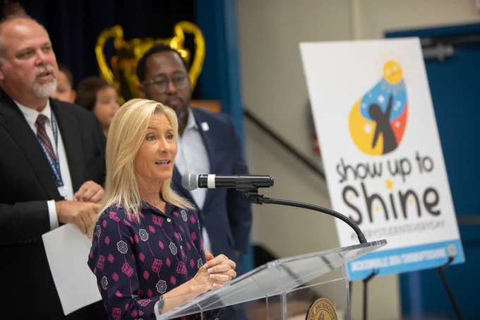 mayor deegan behind a podium speaking with a show up to shine poster in the background