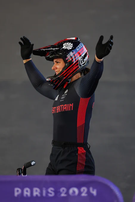 GB rider Beth Shriever lifts her arms and smiles at onlookers (out of shot) whilst wearing Team GB kit at the Paris 2024 Olympic Games. The photo is taken from Beth's left side, with a small portion of a purple 'Paris 2024' barrier visible at the bottom of the image.