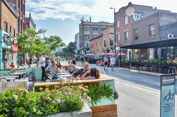 People walk, sit, and dine along a pedestrianized street in Montreal. It is lined with trees, planter boxes, terraces, and benches.